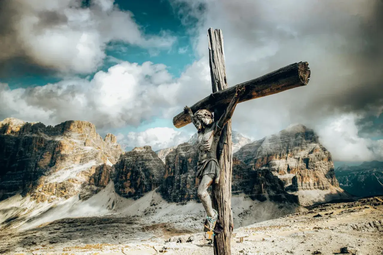 Holzfigur der Jesus-Kreuzigung vor dramatischer Bergkulisse und Wolkenhimmel.