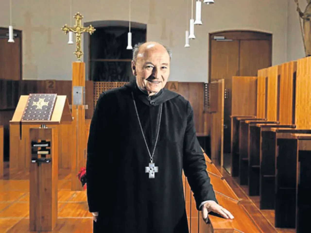 Ein lächelnder Odilo Lechner in schwarzer Soutane mit Kreuzkette steht in einer Kirche vor Beichtstühlen und einem Lesepult.
