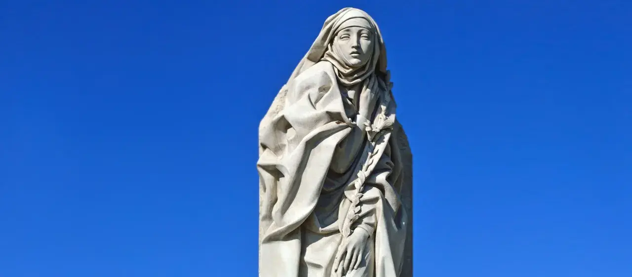 Steinskulptur der Heiligen Katharina von Siena mit Schleier und einem Lorbeerkranz vor blauem Himmel.