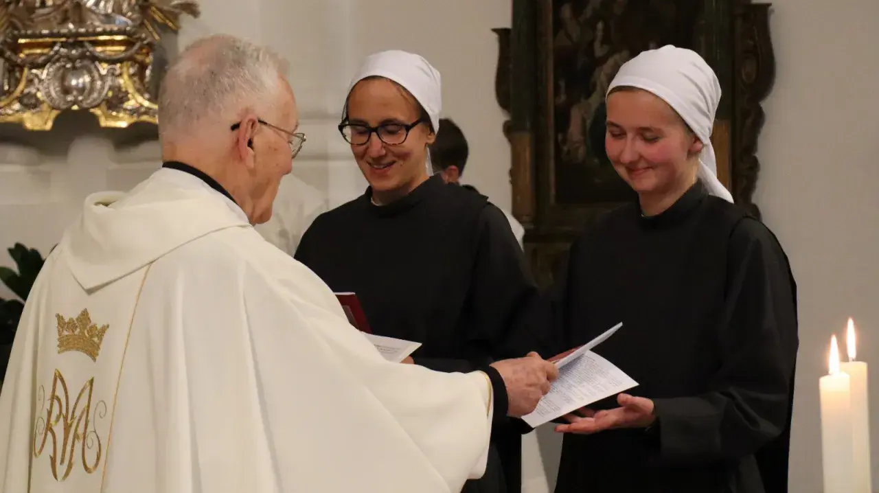 Priester übergibt Dokumente an zwei Dienerinnen vom Heiligen Blut, die lächelnd ihre Aufgaben annehmen.