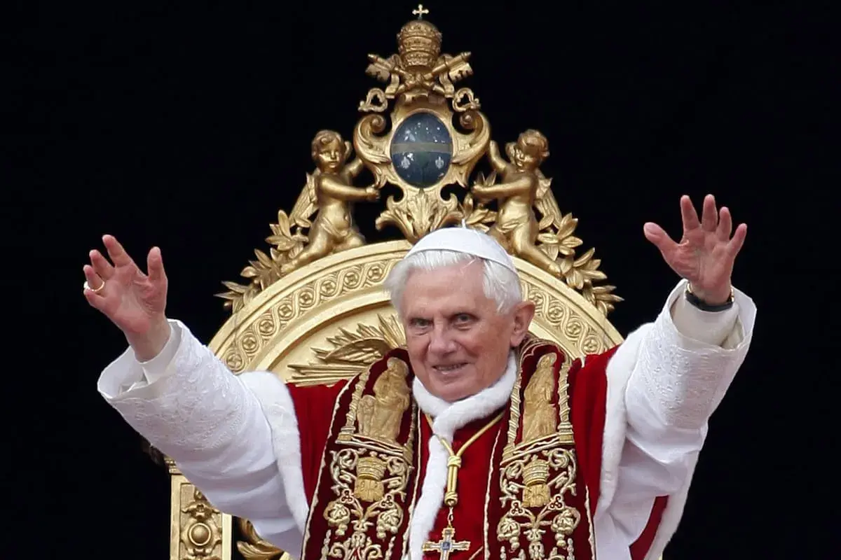 Papst Benedikt XVI. winkt in prächtiger Papst-Kleidung, mit weißer Soutane und rotem Umhang, verziert mit goldenen Stickereien.