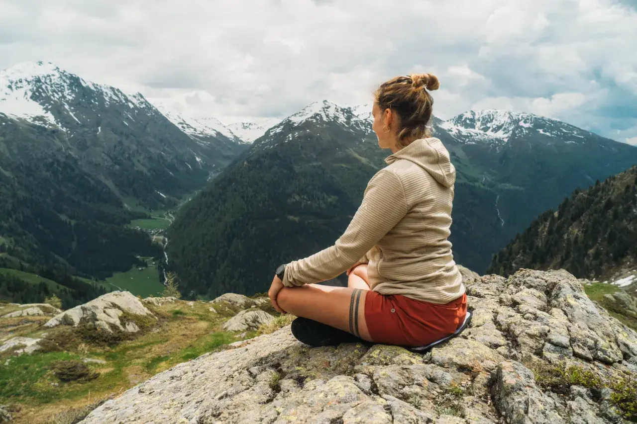 Frau meditiert auf einem Felsen mit Blick auf ein alpines Tal. Sie findet Ruhe und Inspiration für ihre Meditationstexte unterwegs sein.