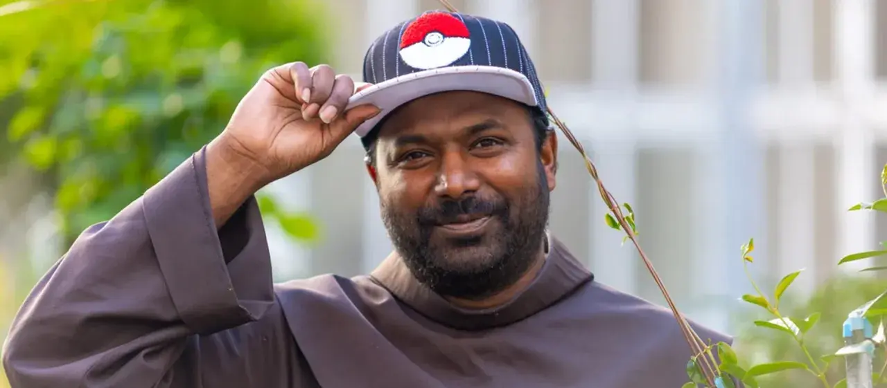 Pater Sandesh Manuel, ein Mann mit Bart, trägt eine Kappe mit Pokémon-Motiv und eine braune Robe. Er lächelt und hebt die Kappe an.