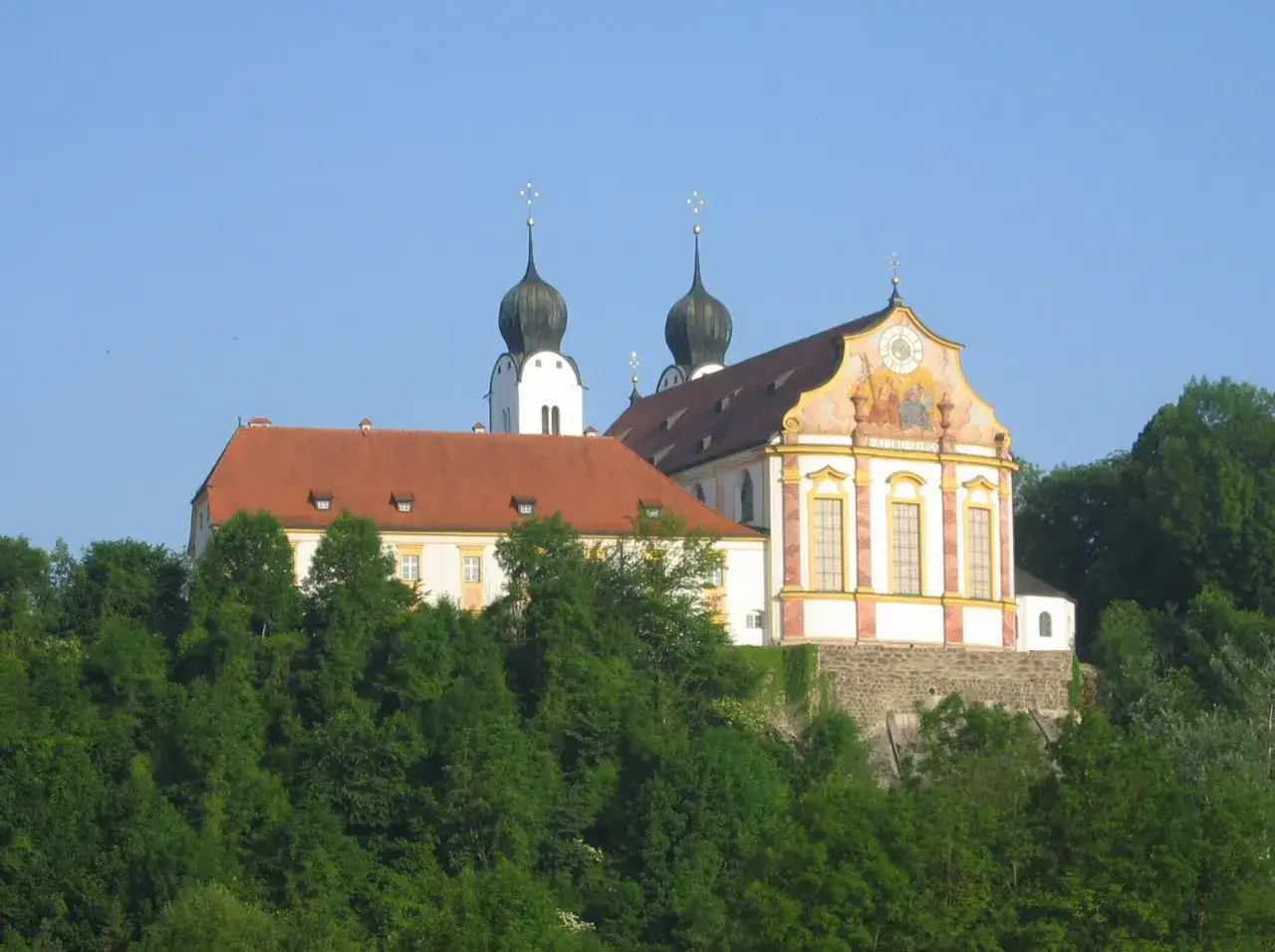 Kloster Baumburg thront auf einem Hügel, umgeben von üppigem Grün. Zwei markante Zwiebeltürme ragen in den blauen Himmel.