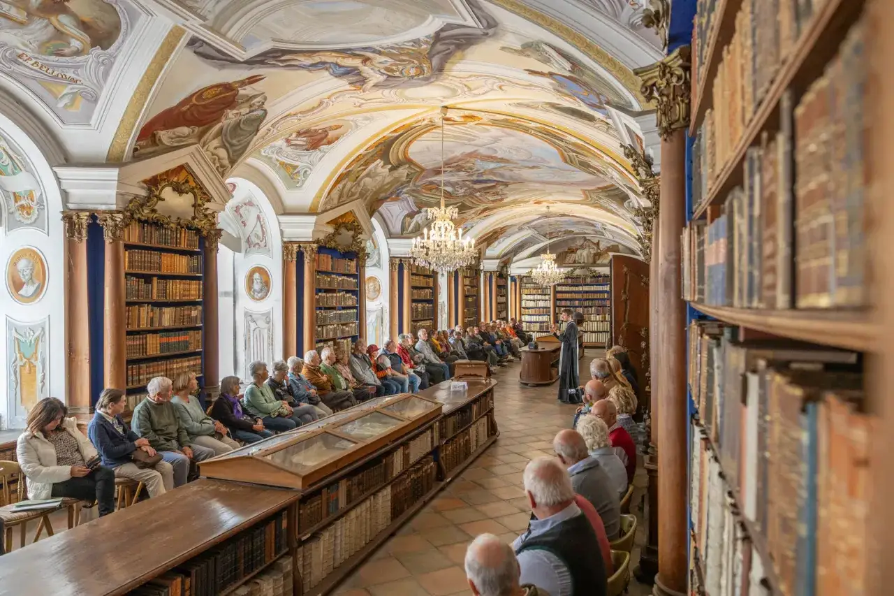 Besucher lauschen einer F&uuml;hrung in der pr&auml;chtigen Bibliothek des Stifts Reichersberg. Deckenmalereien und B&uuml;cherregale s&auml;umen den Raum.