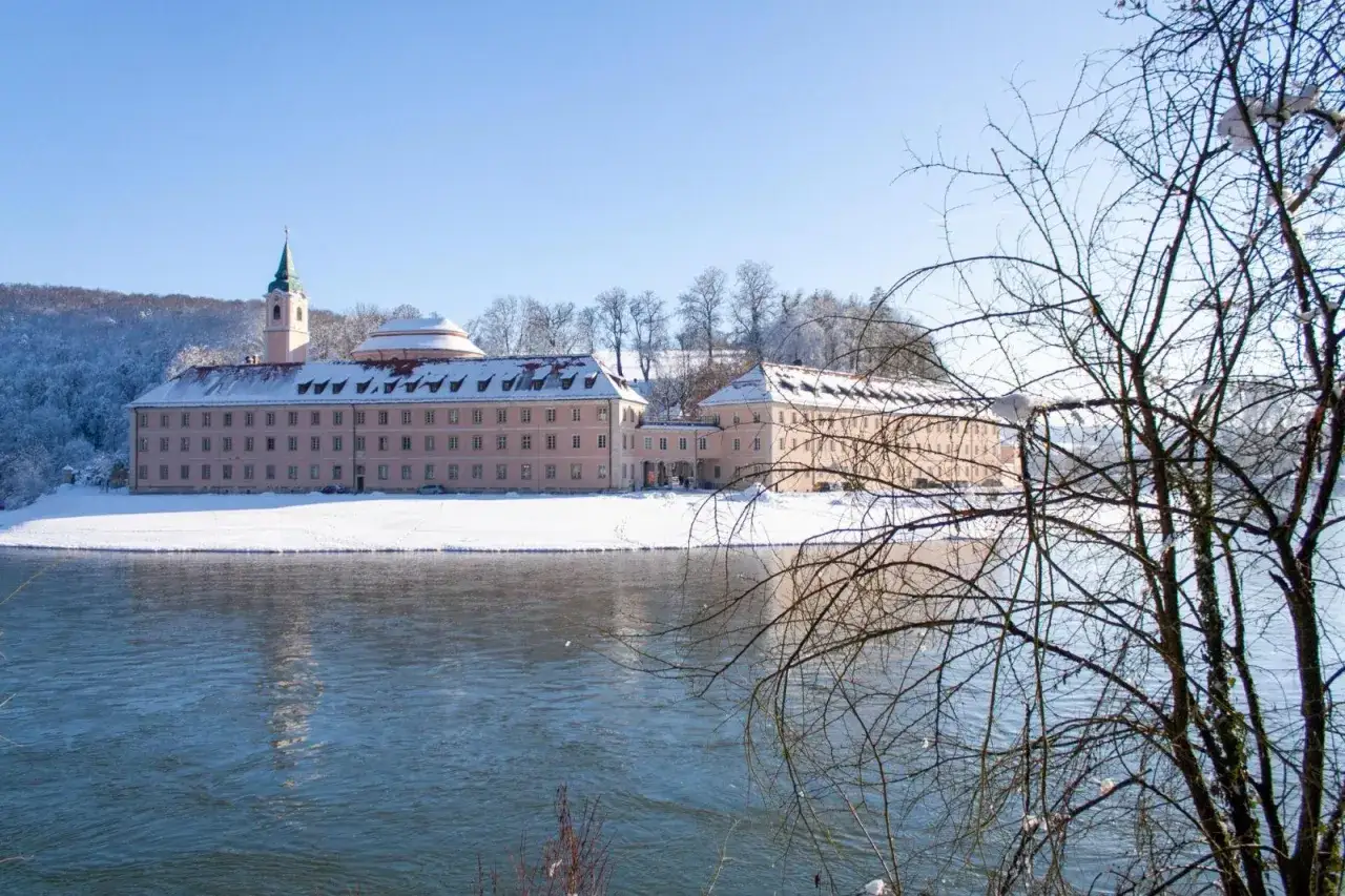 Ein rosa Klostergeb&auml;ude mit Kirchturm am Flussufer im Schnee. Hier k&ouml;nnte man dauerhaft im Kloster leben, umgeben von winterlicher Ruhe.