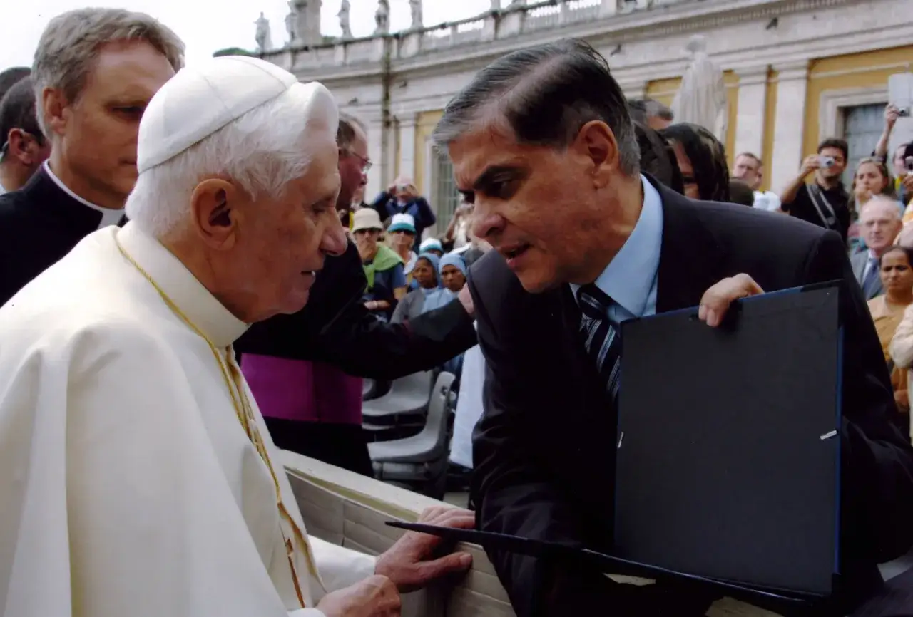 Benedikt XVI. im Gespräch mit einem Mann, der ein schwarzes Mappe hält. Im Hintergrund stehen Menschen.