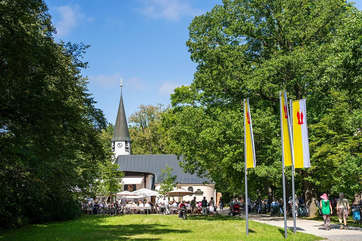 Menschen genießen das Leben vor der Kirche Maria Eich in Planegg. Sonnenschein, grüne Bäume und Fahnen schaffen eine festliche Atmosphäre.