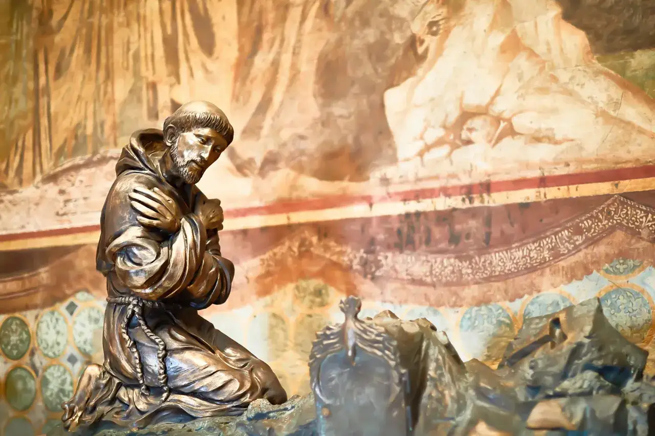 Bronzestatue von St. Franziskus kniend im Gebet vor einer Wandmalerei.