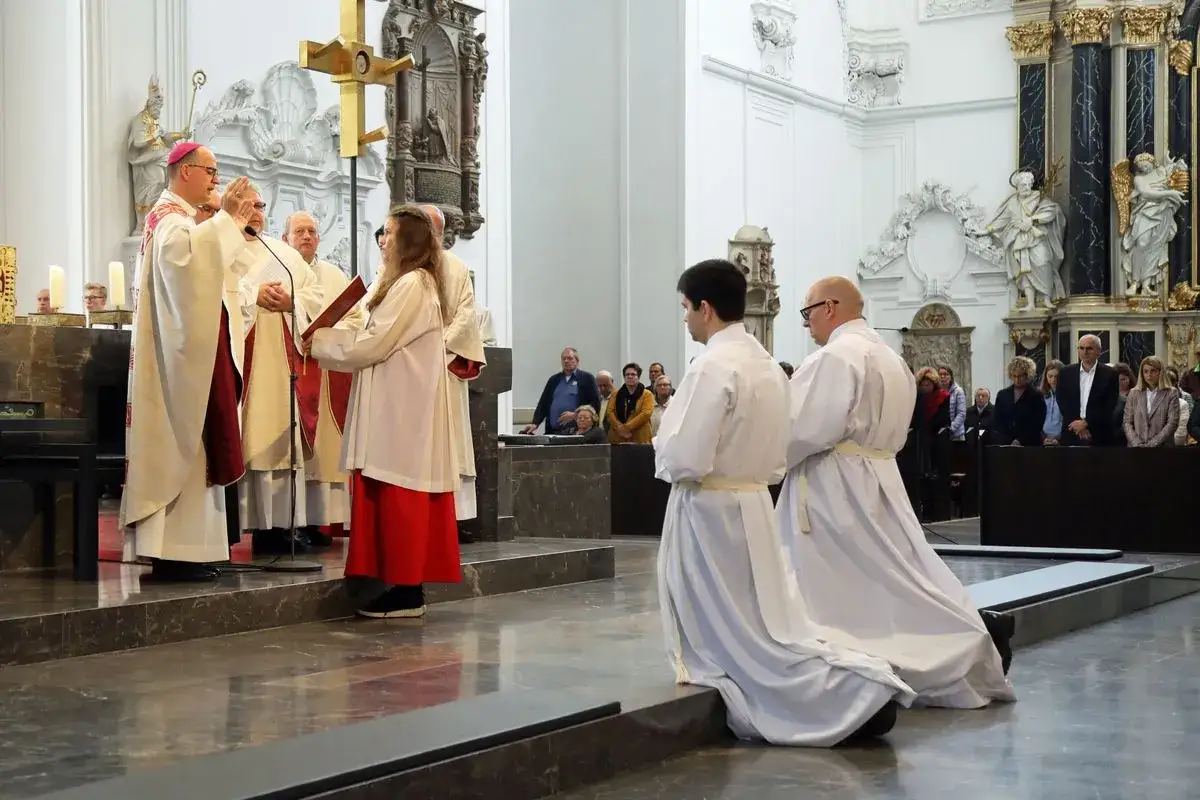 Ein Bischof spendet den Segen. Ministranten in weißen Gewändern knien. Gottes Segen möge sie begleiten.