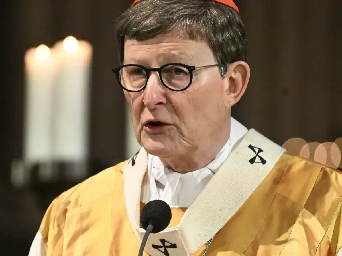 Kardinal Woelki spricht in die Kamera, trägt eine rote Kopfbedeckung und eine goldene Robe. Kerzen sind im Hintergrund zu sehen.