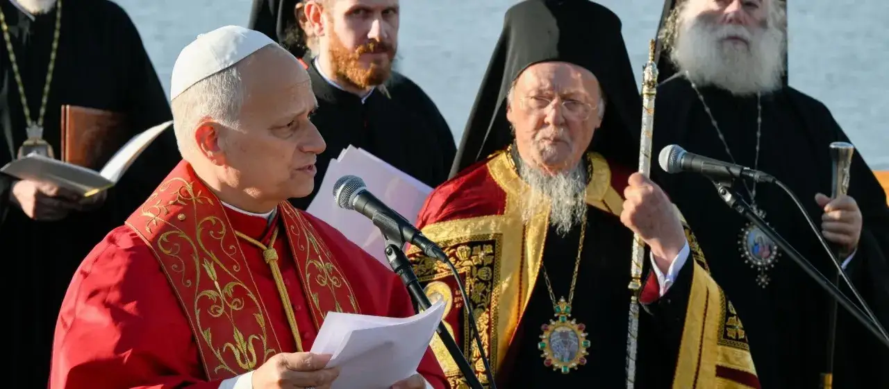 Zwei Geistliche, einer in Rot, sprechen ins Mikrofon. Der andere, ein Patriarch, tr&auml;gt eine Mitra und h&auml;lt einen Stab.