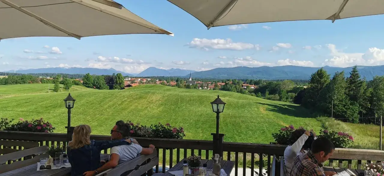 Blick vom Biergarten des Klosters Reutberg auf gr&uuml;ne H&uuml;gel, ein Dorf und die Alpen.