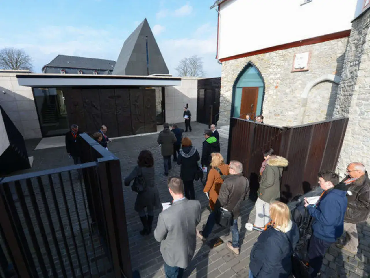 Besucher vor moderner Kapelle mit Pyramidenform. Ein Mann mit orangefarbenem Band, vielleicht der Bischof Limburg, steht in der N&auml;he einer goldenen Badewanne.