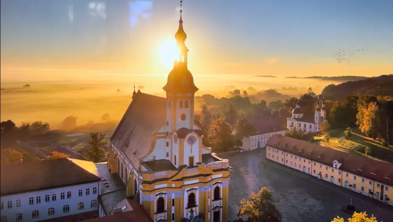 Sonnenaufgang über einem Klosterkomplex, der an die Dienerinnen vom Heiligen Blut erinnert. Nebel liegt über den Hügeln.