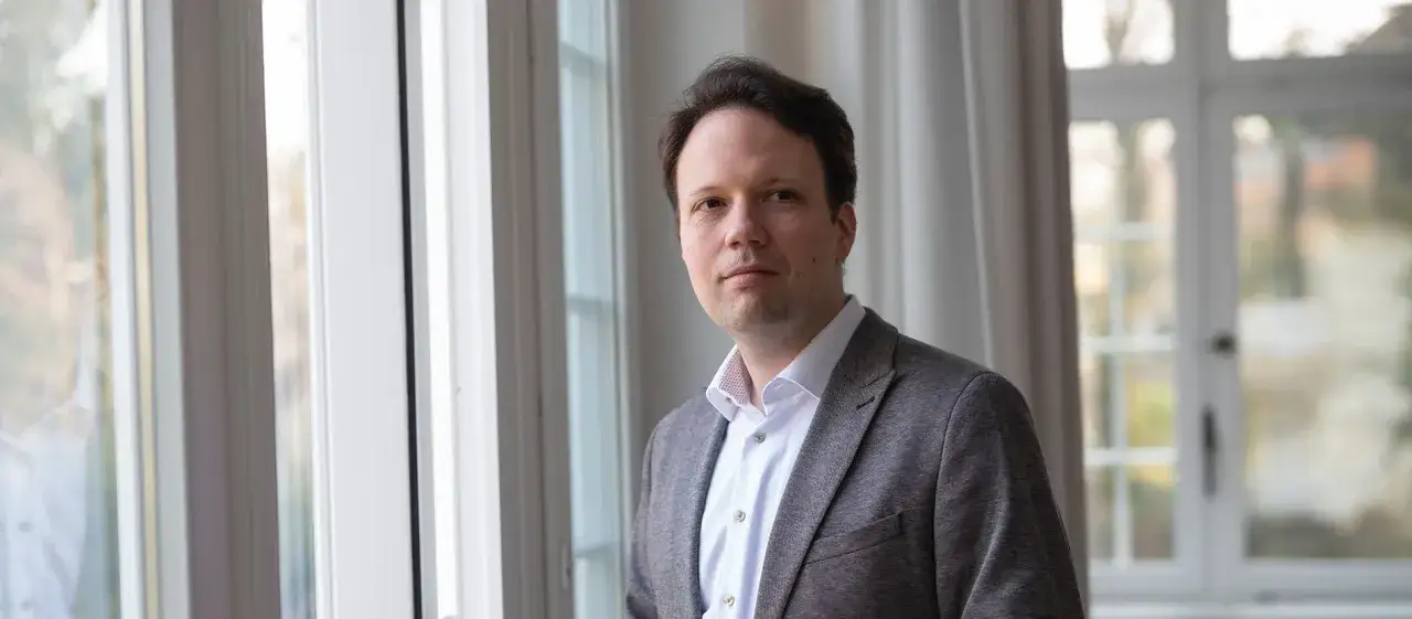 Michael Seewald blickt nachdenklich in die Kamera, im Hintergrund ein Fenster mit Vorh&auml;ngen.