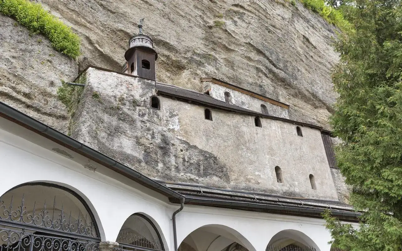 Teil der Erzabtei Salzburg, ein Gebäude schmiegt sich an eine Felswand, mit einem kleinen Turm und Arkaden im Vordergrund.