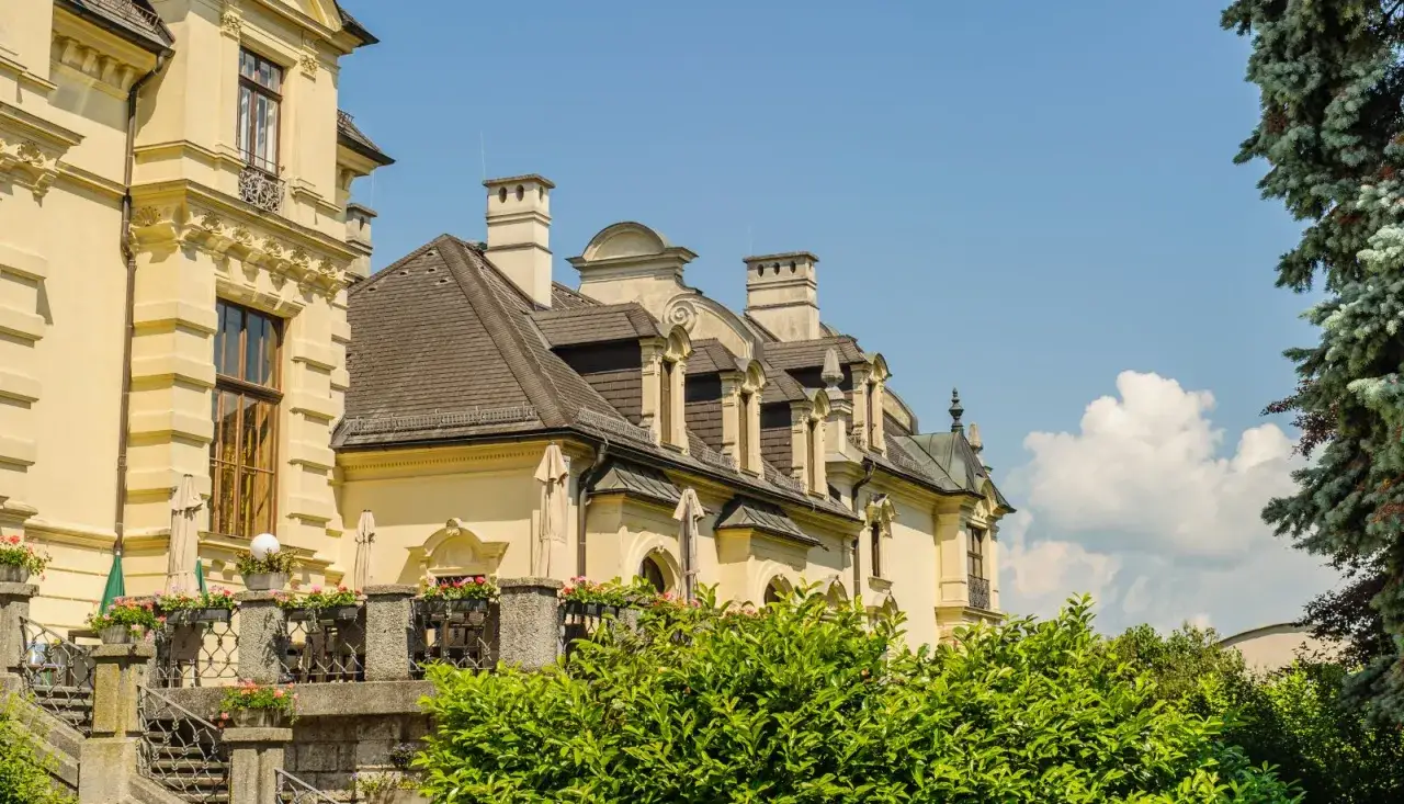 Gelbe Fassade mit detailreicher Architektur und Dachgauben. Ein gr&uuml;ner Baum und bl&uuml;hende Pflanzen umrahmen das Anwesen der Pallottiner Salzburg.