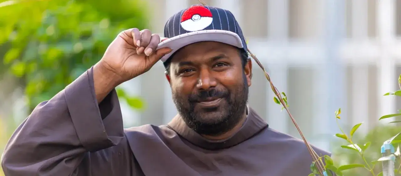 Pater Sandesh Manuel, ein Mann mit Bart, tr&auml;gt eine Kappe mit Pok&eacute;mon-Logo und eine braune Robe.