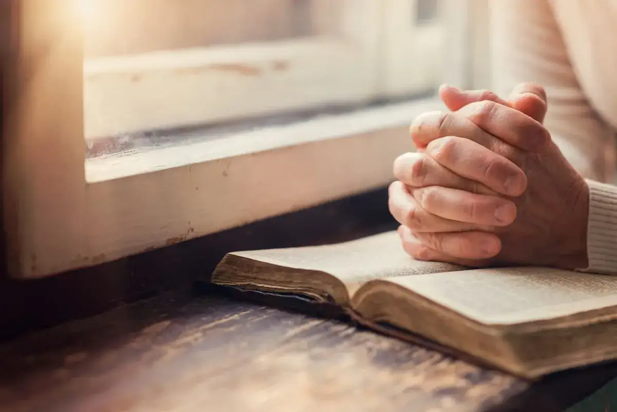 Gefaltete H&auml;nde ruhen auf einer alten Bibel, w&auml;hrend das Licht durch ein Fenster f&auml;llt. Ein Moment der Besinnung, der die Liebe von Gott als Vater widerspiegelt.