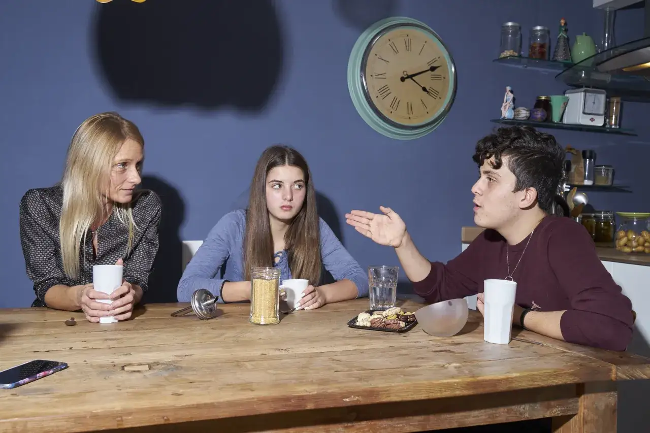 Ein Junge gestikuliert wild, während seine Mutter und Schwester ihn am Tisch ansehen. Ein deutlicher Streit in der Familie liegt in der Luft.