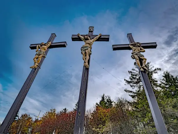 Drei Kreuze mit Jesusfiguren, die die Passion Christi darstellen, vor einem dramatischen Himmel und herbstlichem Laub.