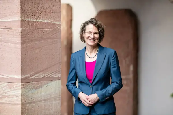 Christiane Tietz, eine Frau mit Brille und lockigem Haar, lächelt in einem blauen Hosenanzug vor einer Sandsteinwand.