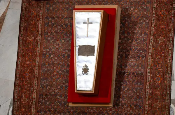 Der gläserne Sarg eines Papstes, mit Kreuz und Wappen, liegt auf einem roten Samtkissen vor einem reich gemusterten Teppich im Petersdom.