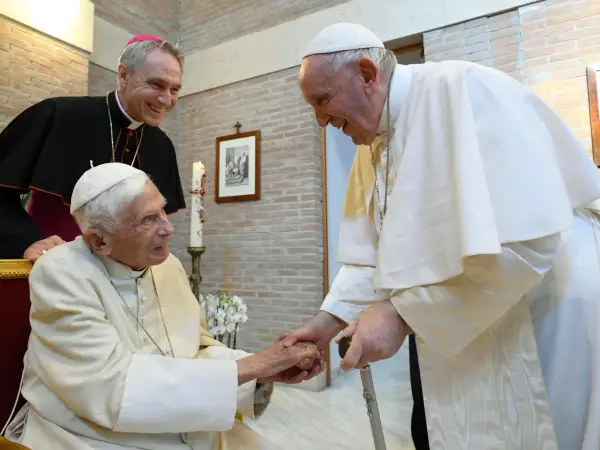 Papst Franziskus und ein Bischof lächeln, während sie Benedikt XVI. die Hand schütteln.
